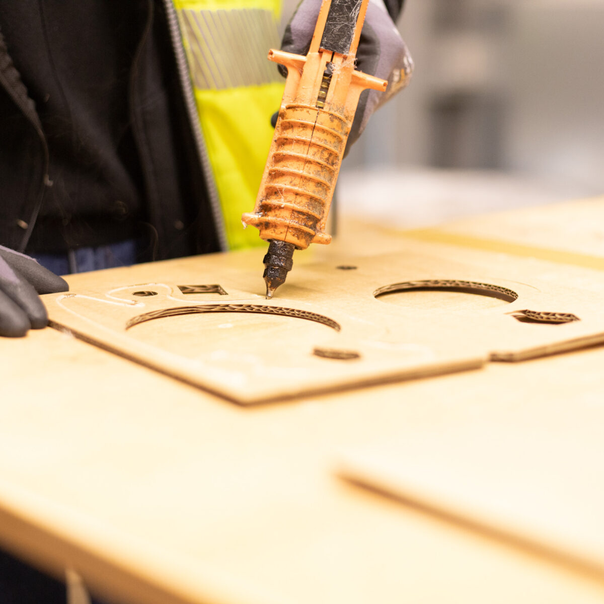 Arbeiter in gelber Sicherheitsweste und Schutzhandschuhen verwendet eine Heißklebepistole auf einem Kartonzuschnitt. Die Person ist konzentriert auf die Anwendung von Heißkleber auf präzise Aussparungen im Karton, wahrscheinlich um Teile für eine Verpackung oder Montage zusammenzufügen. Der Fokus liegt auf der Hand, der Klebepistole und dem Kartonzuschnitt, während der Rest des Bildes unscharf bleibt, was die Handarbeit und den Fertigungsprozess in der Verpackungsindustrie hervorhebt.