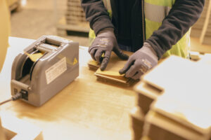 Arbeiter in einer Werkstatt, der Kartonzuschnitte mit einer Maschine bearbeitet. Der Arbeiter trägt eine reflektierende gelbe Sicherheitsweste und Schutzhandschuhe und legt Kartonteile in eine Maschine, die möglicherweise zum Formen oder Schneiden der Pappe dient. Der Fokus liegt auf den Händen des Arbeiters und den Kartonzuschnitten, während die Maschine und die Umgebung im Hintergrund unscharf bleiben, was die Präzisionsarbeit im Verpackungsprozess hervorhebt.
