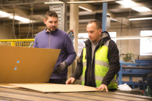 Zwei Arbeiter im Gespräch an einem industriellen Arbeitstisch in einer Fertigungshalle. Der Mann links trägt einen lila Pullover und hält ein großes Stück Karton, während der Mann rechts eine reflektierende gelbe Sicherheitsweste und eine Jacke trägt und auf das Material auf dem Tisch zeigt. Im Hintergrund sind weitere industrielle Ausrüstungen und Materialien zu erkennen. Die Szene zeigt die Zusammenarbeit und Materialprüfung in der Produktion oder Qualitätskontrolle von Tillmann Verpackungen.