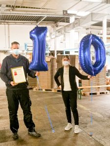 Mitarbeiter steht neben der Geschäftsführerin bekommt zum 10-jährigen Firmenjubiläum eine Urkunde und Helium-Ballons in Form einer 10