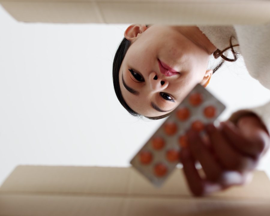 Asiatische Frau blickt in einen Karton und entnimmt einen Tablettenfilmstreifen, Ansicht aus der Froschperspektive des Kartons.