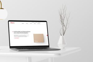 Minimalistisch gestalteter Arbeitsplatz mit einem geöffneten Laptop, der die Webseite von Tillmann Verpackungen anzeigt. Neben dem Laptop steht eine stilvolle Vase mit trockenen Ästen auf einem weißen Tisch. Über dem Tisch hängt eine moderne Lampe. Diese Szenerie vermittelt ein professionelles, sauberes und modernes Image, welches gut zu einem Unternehmen passt, das Wert auf Design und Präsentation legt.