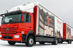 Lkw von Tillmann Verpackungen im Einsatz bei winterlichen Bedingungen. Der rote Mercedes-Benz Lkw trägt auf seiner Plane groß das Firmenlogo von Tillmann mit dem Slogan "GUT VERPACKT" sowie Bilder, die das Verpacken von Produkten darstellen.