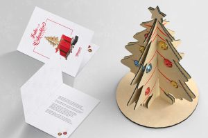Weihnachtliche Grußkarte und Dekorationsbaum aus Karton von Tillmann Verpackungen. Links im Bild ist eine aufgeklappte Grußkarte mit den Worten "Fröhliche Weihnachten" und einem ausgestanzten Tannenbaum-Motiv, das teilweise in 3D hervortritt. Rechts ist ein dreidimensionaler, aufstellbarer Karton-Weihnachtsbaum mit eingesetzten, farbigen Kugeln dargestellt.