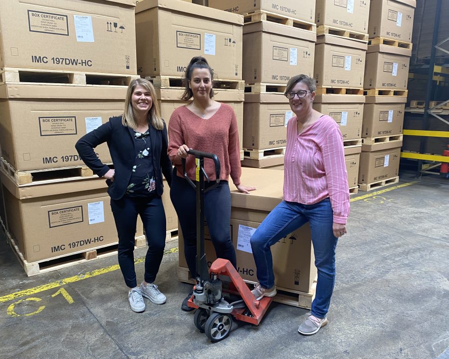 Drei lächelnde Frauen in einem Lagerhaus mit gestapelten Kartons im Hintergrund. Eine Frau steht links mit verschränkten Armen, eine steht in der Mitte und lehnt sich an einen Handhubwagen, und die dritte Frau steht rechts und stützt sich auf einen der Kartons. Die Kartons sind groß und tragen Etiketten, die auf industrielle oder kommerzielle Waren hinweisen könnten. Die Umgebung zeigt ein typisches Lagerhaus-Szenario mit gelber Bodenmarkierung für Sicherheitszonen.