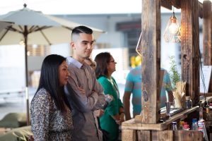 Das Foto zeigt zwei Personen, die an einem Event teilnehmen. Eine asiatische Frau und ein Mann mit einem gepflegten Haarschnitt stehen nebeneinander an einer Bar und warten auf Ihre Bestellung.