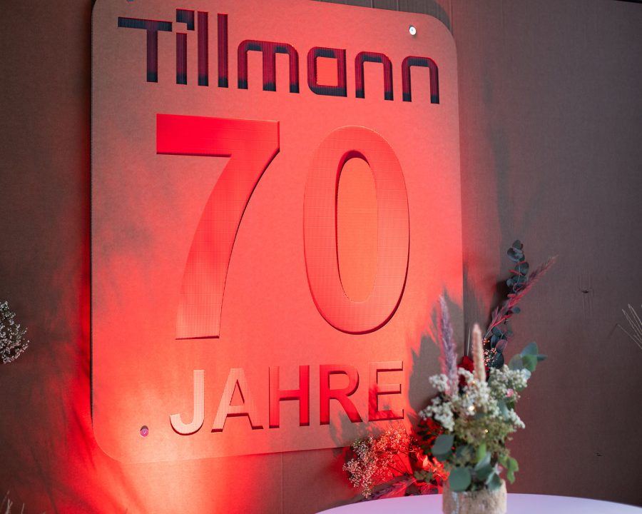 Feierliche Anzeige zum 70-jährigen Jubiläum von Tillmann Verpackungen. Ein großes Schild mit der Aufschrift "Tillmann 70 Jahre" in roter Beleuchtung, vor einem dunklen Hintergrund, mit Blumenarrangements im Vordergrund. Die Beleuchtung und die Blumendekoration verleihen der Szene ein festliches Ambiente, was auf eine Jubiläumsfeier oder ein Unternehmensereignis hindeutet.