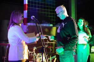 Eine Frau und ein Mann stehen auf einer beleuchteten Bühne bei einer Veranstaltung, die Frau liest etwas von einem Blatt Papier ab und übergibt es dem Mann. Beide lächeln und scheinen in einer angenehmen Interaktion zu sein. Im Hintergrund ist eine weitere Person mit einem Mikrofon zu sehen.