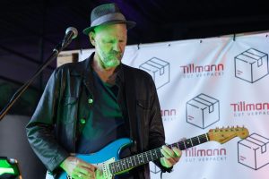 Das Bild zeigt einen Gitarristen, der konzentriert auf seiner blauen E-Gitarre spielt. Er trägt einen schwarzen Hut und eine schwarze Jacke.