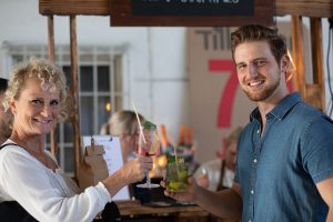 Eine ältere Frau und ein jüngerer Mann stoßen mit Cocktails an einem Veranstaltungsort an. Sie lächeln und sehen entspannt aus, möglicherweise im Rahmen einer Firmenveranstaltung oder Feierlichkeit. Im Hintergrund ist deutlich ein Schild mit der Aufschrift "Tillmann 70 Jahre" zu erkennen.