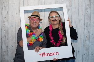 Zwei Personen, die Spaß haben und mit einem Rahmen posieren, auf dem "Tillmann" steht. Sie tragen festliche Accessoires: Der Mann links trägt einen Strohhut, eine bunte Blumenkette und eine lustige Brille, während die Frau rechts ebenfalls eine Brille und einen Schnurrbart an Stäben sowie eine rosa Blumenkette trägt.