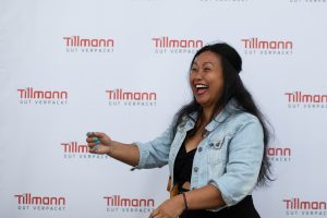 Eine fröhlich lachende Frau steht vor einem Hintergrund mit dem Tillmann Verpackungen Logo. Sie trägt eine Jeansjacke und gestikuliert mit der Hand, was auf eine lebhafte Interaktion oder einen Moment der Freude während einer Veranstaltung hindeutet. Der Hintergrund dient als Fotowand, typisch für Events, wo Gäste fotografiert werden.