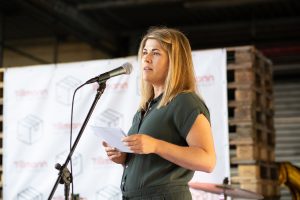Kathrin Hildebrand spricht in ein Mikrofon und hält eine Rede. Sie steht vor einem Hintergrund, der das Logo von Tillmann Verpackungen zeigt. Im Hintergrund sind auch Paletten zu sehen, was darauf hindeutet, dass die Veranstaltung in einem Lager oder industriellen Umfeld stattfindet.