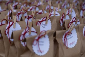 Eine Serie von braunen Papiergeschenktüten, die sorgfältig mit individuellen Namensschildern und rot-weißen Schnüren versehen sind. Jedes Namensschild ist mit einer Wäscheklammer an der Tüte befestigt. Die Namensschilder haben ein dekoratives Muster am Rand und zeigen verschiedene Namen, was darauf hindeutet, dass diese Tüten für Gäste oder Teilnehmer einer Veranstaltung vorbereitet wurden.