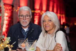 Ein älteres Paar genießt scheinbar eine festliche Veranstaltung. Beide halten Cocktails und lächeln in die Kamera. Hinter ihnen ist eine warme Beleuchtung zu sehen, die für eine gemütliche und festliche Stimmung sorgt. Ihre entspannten Gesichtsausdrücke und die festliche Atmosphäre deuten auf eine gesellige Zusammenkunft hin.