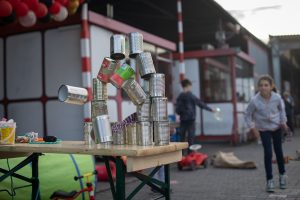 Ein Spieltisch mit einer Pyramide aus Konservendosen, die als Ziel für ein Wurfspiel auf einem Fest aufgestellt wurden. Im Hintergrund sind Kinder zu sehen, die spielen und Spaß haben. Die Szene befindet sich im Freien, erkennbar an der Tageslichtbeleuchtung und den Gebäuden im Hintergrund.