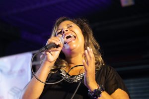 Eine Sängerin performt leidenschaftlich in ein Mikrofon, ihre Augen sind geschlossen und ihre Handgestik unterstreicht die Intensität ihrer Darbietung. Der Hintergrund ist in blaues Licht getaucht, was auf eine Bühne mit Beleuchtung hinweist. Sie scheint vollkommen in ihrer Musik aufzugehen, was typisch für eine Live-Performance auf einem Event oder Konzert ist.