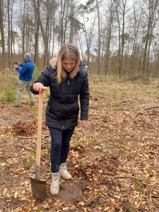 Das Bild zeigt Kathrin Hildebrand, die Geschäftsführerin von Tillmann Verpackungen, wie sie in einem deutschen Wald einen Sprössling pflanzt. Im Hintergrund sind viele Bäume zu sehen sowie ein Mann mit Blauer Fleecejacke der scheinbar ein Foto schießt.