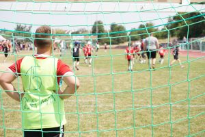 Fußballspiel des Tillmann Cups auf einem sonnigen Fußballplatz mit Kindern in verschiedenen Trikots. Im Hintergrund sind Zuschauer und Bäume zu erkennen.